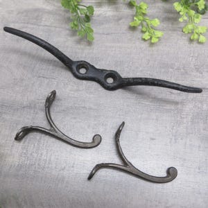 May include: Three dark metal hooks. One is a long, curved bar with two holes, and the other two are smaller, Y-shaped hooks. The hooks are likely for hanging items and have a rustic, antique appearance.