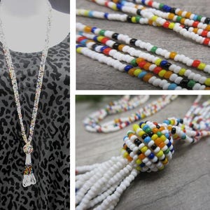 May include: A long beaded necklace with a white base and colorful accents. The necklace features a tassel pendant with a beaded ball and a fringe of white beads. The beads are small and round, with a mix of colors.