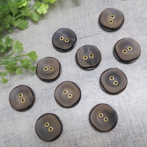 May include: A collection of ten dark brown wooden buttons with two gold-colored holes each. The buttons have a slightly irregular, organic shape and are arranged on a light gray surface. The buttons are a craft or sewing supply.
