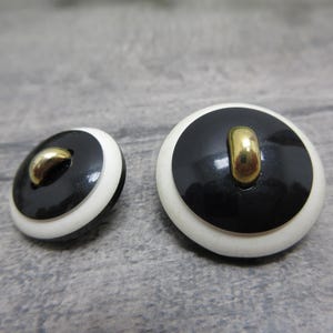 May include: Two vintage buttons with a black center, white border, and gold-colored loop. The buttons are round and have a glossy finish. One button is slightly smaller than the other. The buttons are likely for clothing or crafts.