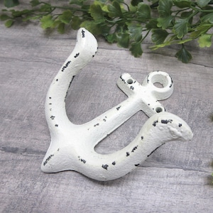 May include: A white wall hook shaped like a nautical anchor with a distressed finish showing black metal underneath. The hook features two curved arms for hanging items and a circular mounting point at the top with two pre-drilled screw holes.