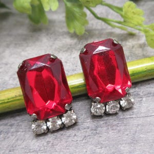 May include: A pair of red gemstone earrings with a silver setting. The gemstones are rectangular and have a faceted cut. The earrings have a cluster of smaller clear gemstones around the base of the larger red gemstone.
