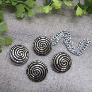 May include: Four silver-toned buttons with a spiral design and a silver chain. The buttons are round and have a raised spiral pattern. The chain is made of small, interlocking links. The buttons and chain are on a gray surface.