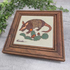 May include: A square ceramic tile featuring a brown armadillo illustration above a green prickly pear cactus with orange flowers, set in a dark brown wooden frame. The artist signature Teissedre is visible in the bottom left corner of the tile.