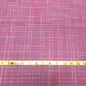 May include: A close-up of a pink fabric with a multi-coloured plaid pattern. A yellow measuring tape is laid across the fabric, showing measurements from 1 to 15 inches.