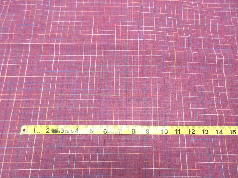 May include: A close-up of a pink fabric with a multi-coloured plaid pattern. A yellow measuring tape is laid across the fabric, showing measurements from 1 to 15 inches.