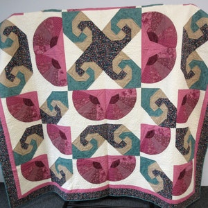 May include: A quilt featuring a repeating geometric pattern of cream, teal, tan, and dark floral fabric blocks. The design includes semicircular fan shapes and angular, swirling motifs arranged in a grid, finished with a dark floral border.