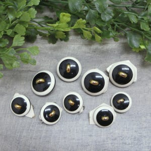 May include: A collection of vintage black and white buttons with gold-colored accents. The buttons have a circular design with a black center, a white border, and a gold-colored bar in the center. Some buttons have attached white fabric.