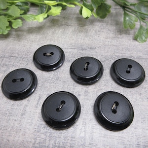 May include: Six black, round buttons with a glossy finish are arranged on a gray surface. Each button has a recessed center with either two holes or a metal shank for sewing. Green foliage is visible in the background.