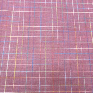 May include: A close-up of a red fabric with a multi-coloured plaid pattern. The fabric has a subtle texture and a soft, woven appearance.