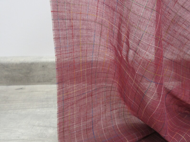 May include: A close-up of a red fabric with a checkered pattern. The fabric has thin white, yellow, and blue lines woven into the red fabric.