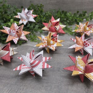 May include: A collection of eight three-dimensional paper stars featuring various patterns in shades of red, orange, yellow, and white. Each star is constructed from folded paper segments, creating a multi-pointed geometric shape with intricate designs.