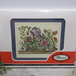 May include: A Pimpernel De Luxe casserole stand in its original white packaging with a red accent band. The product features a rectangular floral illustration of a colorful bouquet in a basket, framed by a dark blue border on a cream background.