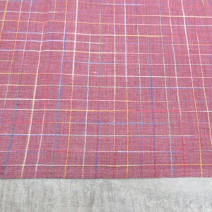 May include: A close-up of a red fabric with a multi-coloured plaid pattern. The fabric has a subtle texture and a slightly faded appearance.