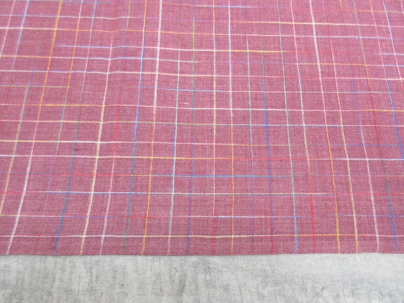 May include: A close-up of a red fabric with a multi-coloured plaid pattern. The fabric has a subtle texture and a slightly faded appearance.