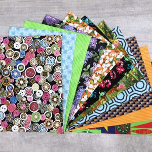 May include: A collection of patterned fabric squares fanned out on a gray surface. The fabrics feature various designs, including buttons, floral patterns, stripes, and geometric shapes. Colors include black, pink, blue, green, orange, and white.