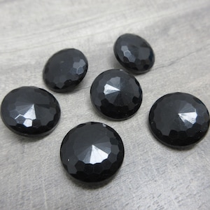 May include: Six faceted black buttons with a glossy finish are scattered on a light gray surface. The buttons have a round shape with a geometric pattern, reflecting light. These buttons are suitable for sewing and craft projects.