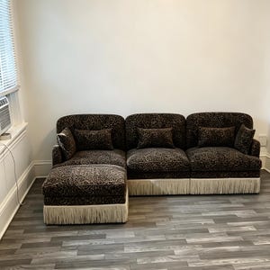 May include: A modular sectional sofa upholstered in a brown and black leopard print fabric. The sofa features three main seating sections, a chaise lounge extension, and matching rectangular throw pillows. The base is finished with cream fringe.