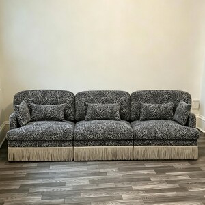 May include: A three-section modular sofa upholstered in a black and white leopard print fabric. The sofa features rounded backrests, three rectangular throw pillows, and a base skirt finished with long, light beige fringe trim along the bottom.