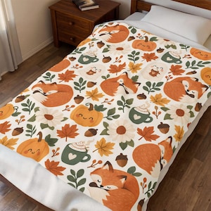 May include: A white fleece blanket featuring a repeating pattern of sleeping orange foxes, smiling pumpkins, green mugs with whipped cream, white daisies, brown acorns, and orange maple leaves spread across the fabric surface.