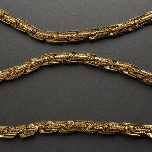 May include: A gold-toned chain necklace featuring a complex, interlocking link design with a textured, reflective surface. The chain is arranged in three horizontal, parallel rows, showcasing the repeating geometric pattern of the metal links.