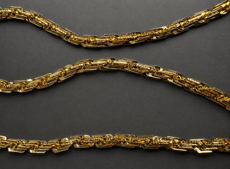May include: A gold-toned chain necklace featuring a complex, interlocking link design with a textured, reflective surface. The chain is arranged in three horizontal, parallel rows, showcasing the repeating geometric pattern of the metal links.