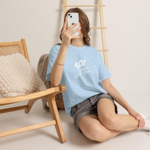 Op de afbeelding: Een lichtblauw t-shirt met korte mouwen en de witte tekst "SQL Secure Queen Lives" op de borst. Het shirt heeft een ronde hals en een relaxte pasvorm, gemaakt van een effen gekleurde stof met een gladde, matte afwerking.
