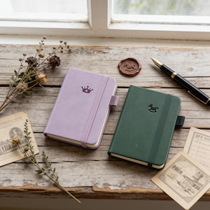 May include: A light purple notebook with a crown icon and a dark green notebook with a rocking horse icon, both featuring elastic closures and pen loops, accompanied by a fountain pen, a wax seal, and dried botanical elements on a wooden surface.