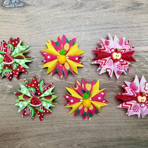 May include: Five star-shaped hair bows made of folded ribbon. Two feature red, green, and white watermelon patterns with watermelon slice centers. Two feature yellow and pink pineapple patterns with pineapple centers. One features pink apple patterns with an apple center.