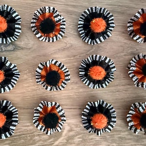 Op de afbeelding: Twaalf ronde rozetten gerangschikt in een raster, elk met geplooide zwart-wit gestreepte stoffen randen, een glinsterende oranje tussenlaag en een centrale pluizige pompon in effen zwart of feloranje.
