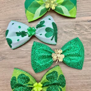 Op de afbeelding: Vier haarstrikken met groene en witte patronen. Eén strik heeft een groen ruitjespatroon met een klavertje van lovertjes. Een andere is wit met groene glitterklavertjes. Een derde is effen groen met een bloemenpatroon en een gouden klavertje. Eén is groen met glitterklavertjes en een limoengroen klavertje.