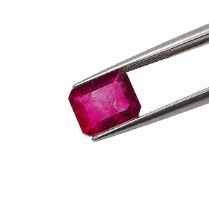 K&ouml;nnte beinhalten: Ein leuchtender, rechteckig geschliffener Rubin mit tiefroter Farbe und sichtbaren inneren Einschl&uuml;ssen, der sicher von einer silberfarbenen Metallpinzette vor einem schlichten wei&szlig;en Hintergrund gehalten wird und seine facettierte Oberfl&auml;che zeigt.