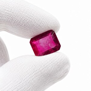 K&ouml;nnte beinhalten: Ein rechteckiger, facettierter Edelstein in einer tiefen, leuchtenden magentaroten Farbe. Der Stein weist scharfe, klare Kanten und eine polierte Oberfl&auml;che auf, die das Licht reflektiert und eine innere Klarheit sowie einen satten Farbton zeigt.