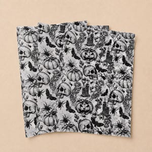 May include: A white fabric featuring a repeating black ink illustration pattern of skulls, pumpkins, spiders, moths, candles, and skeletal hands. The detailed line art creates a dense, monochromatic design across the entire surface of the material.