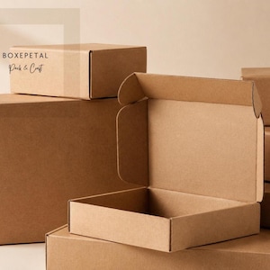 Puede incluir: Una colección de cajas de envío de cartón marrón de varios tamaños, incluida una caja abierta con tapa abatible. La caja cerrada superior presenta el texto impreso BOXEPETAL Pack & Craft en tinta negra en su lateral.
