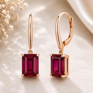 Peut inclure: Une paire de boucles d'oreilles pendantes en or rose avec des pierres précieuses rouge foncé de taille émeraude rectangulaire. Chaque boucle est suspendue à une attache dormeuse incurvée, avec une monture en métal poli fixant les pierres.