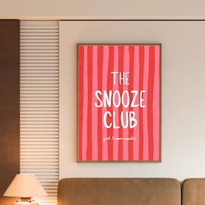May include: A rectangular wall art print featuring vertical stripes in alternating shades of red and pink. The text in white reads THE SNOOZE CLUB and just 5 more minutes in a casual, handwritten font style, centred within a thin wooden frame.