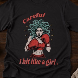 May include: A black t-shirt featuring a graphic of a figure with snakes for hair wearing red boxing gloves. The text "Careful" is printed in red above the figure, and the text "I hit like a girl." is printed in red below the figure.