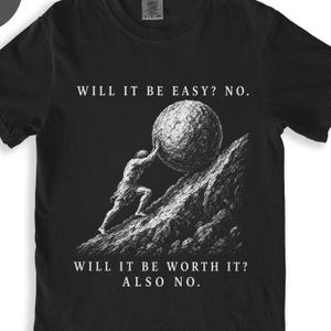 May include: A black short-sleeved t-shirt featuring a white graphic of a figure pushing a large boulder up a rocky hill. The text above reads WILL IT BE EASY? NO. and the text below reads WILL IT BE WORTH IT? ALSO NO. The shirt has a crew neckline.