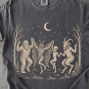 May include: A charcoal gray t-shirt featuring a beige graphic print of a Bigfoot, a moth-like creature, an alien, and a goat-like figure holding hands in a circle. The scene includes trees, mushrooms, a crescent moon, and stars in the background.