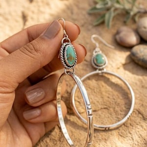 May include: A pair of silver hoop earrings featuring a turquoise stone set in a twisted rope bezel. The earrings have a hook-style closure and a smooth silver hoop with small etched rectangular details along the bottom edge of the metal frame.