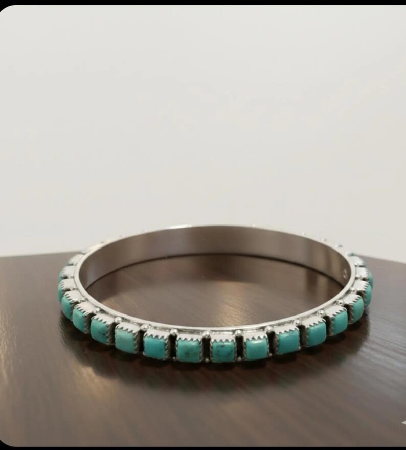 Puede incluir: Un brazalete de plata con una hilera continua de peque&ntilde;as piedras de turquesa de corte cuadrado engastadas en biseles met&aacute;licos individuales. La pulsera tiene un acabado met&aacute;lico pulido y una forma circular uniforme con un patr&oacute;n repetitivo.