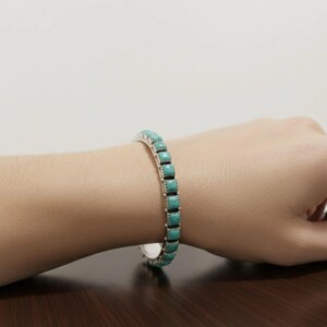 Puede incluir: Una pulsera de plata tipo brazalete con una hilera continua de piedras turquesa cuadradas de color azul claro. El engaste de metal tiene una textura de cuentas en los bordes y la pulsera es una banda r&iacute;gida abierta que rodea la mu&ntilde;eca.