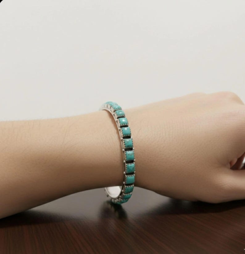 Puede incluir: Una pulsera de plata tipo brazalete con una hilera continua de piedras turquesa cuadradas de color azul claro. El engaste de metal tiene una textura de cuentas en los bordes y la pulsera es una banda r&iacute;gida abierta que rodea la mu&ntilde;eca.