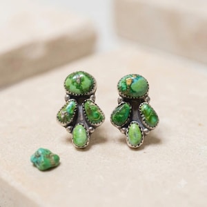May include: A pair of stud earrings featuring clusters of four oval-shaped green turquoise stones set in silver. Each earring has one larger stone at the top and three smaller stones arranged below. A single loose green turquoise stone sits nearby.