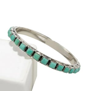 Puede incluir: Una pulsera tipo brazalete de plata con una hilera continua de peque&ntilde;as piedras de turquesa de corte cuadrado engastadas en biseles met&aacute;licos individuales. La pulsera tiene un acabado met&aacute;lico pulido y una banda interior lisa y redondeada.