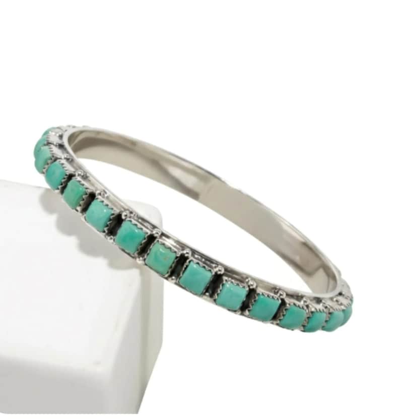 Puede incluir: Una pulsera tipo brazalete de plata con una hilera continua de peque&ntilde;as piedras de turquesa de corte cuadrado engastadas en biseles met&aacute;licos individuales. La pulsera tiene un acabado met&aacute;lico pulido y una banda interior lisa y redondeada.