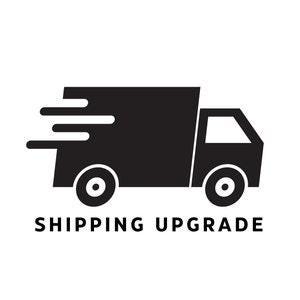 Puede incluir: Un icono gráfico negro de un camión de reparto orientado a la derecha con tres líneas de movimiento horizontales en la parte trasera. Debajo del camión, el texto dice SHIPPING UPGRADE en una fuente negra, negrita y sin remates.