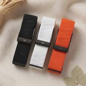 May include: Three watch bands in black, white, and orange, each featuring a textured surface and a matte black metal buckle with a small white letter W logo. The bands are arranged vertically, showcasing a consistent design with a secure clasp.