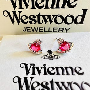 Peut inclure: Une paire de boucles d'oreilles argentées avec une pierre centrale rouge en forme de cœur entourée d'un anneau métallique, évoquant une planète. Elles sont présentées sur une carte blanche avec le texte noir Vivienne Westwood Jewellery.